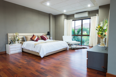 a large room with a hardwood floor, a tall window overlooking trees, a two-person bed with a single bedside table, anda  padded chair next to the window